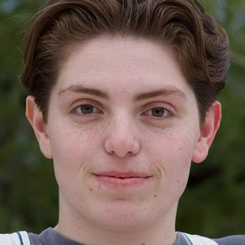 A young person with short brown hair and fair skin is facing the camera outdoors. They have light-colored eyes, freckles, and a slight smile. The background is blurry greenery, indicating an outdoor setting.