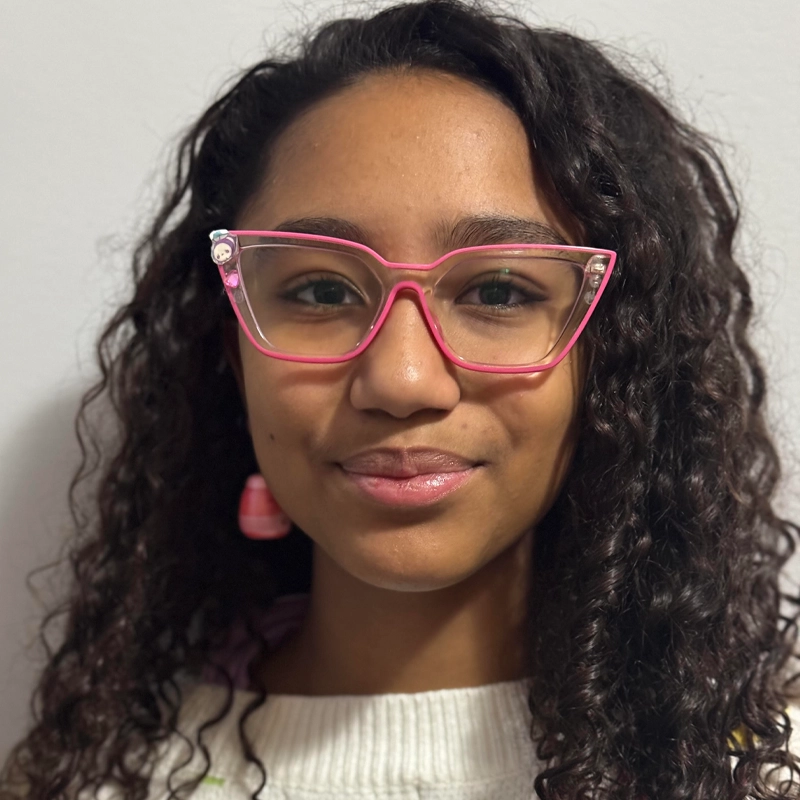 Misa Socorro, a young woman with long, curly dark hair, wears large pink cat-eye glasses and a white sweater. She smiles slightly, looking directly at the camera against a plain white background, with pink earrings as accents.