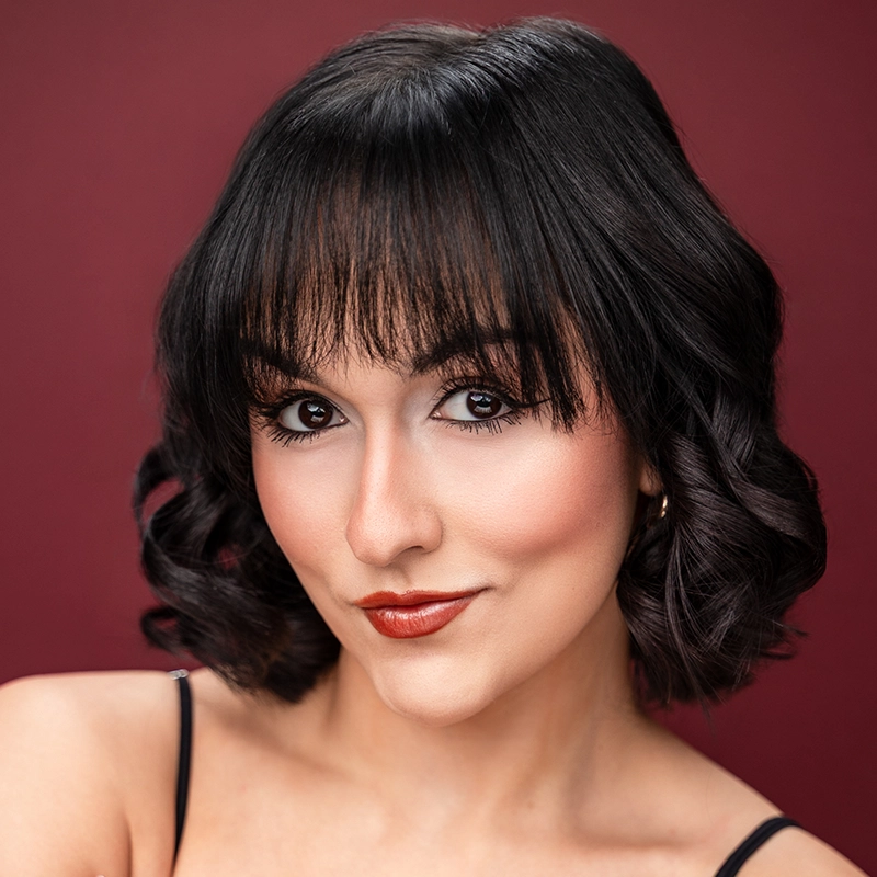 A woman with wavy dark hair and bangs smiles slightly against a burgundy background. She has brown eyes, winged eyeliner, and wears red lipstick. Dressed in a black spaghetti strap top, Sara Ross Avery faces the camera closely.