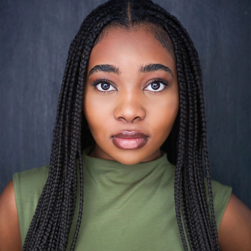 Symoné Spencer, a young woman with long, dark braid extensions, stares directly at the camera. She has smooth brown skin, full lips, and expressive eyes. She wears a sleeveless green top and stands against a dark, textured background.