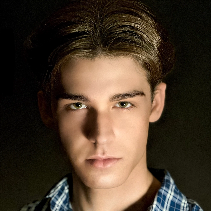 A young man with fair skin, hazel eyes, and short brown hair parted in the middle stares directly at the camera with a neutral expression. Addison Zacharias wears a blue plaid shirt against a dark background that draws focus to his face.