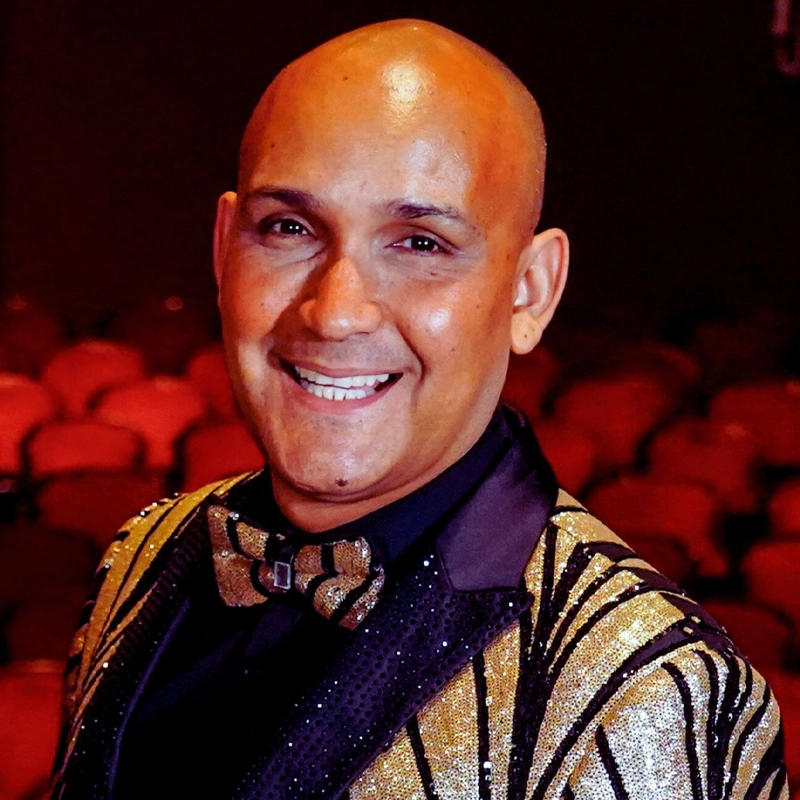 A smiling bald man, Ricardo Stevan, poses in a dark theater with red seats. He wears a glittery gold and black jacket, a matching bow tie, and a black shirt. The softly blurred background highlights his cheerful expression and festive look.