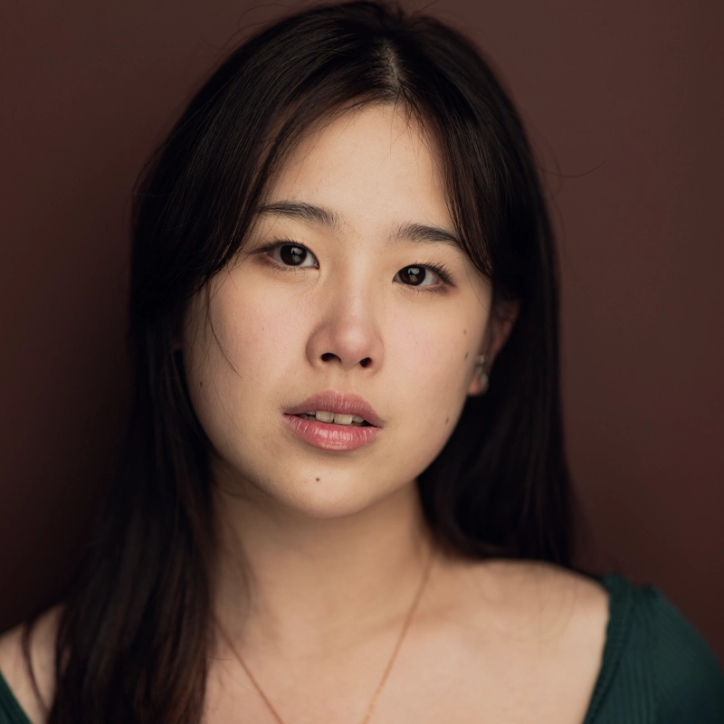 A young woman with long dark hair and fair skin faces the camera against a plain maroon background. She has a neutral expression, wearing a green top and a delicate necklace. The lighting is soft, highlighting her natural features.