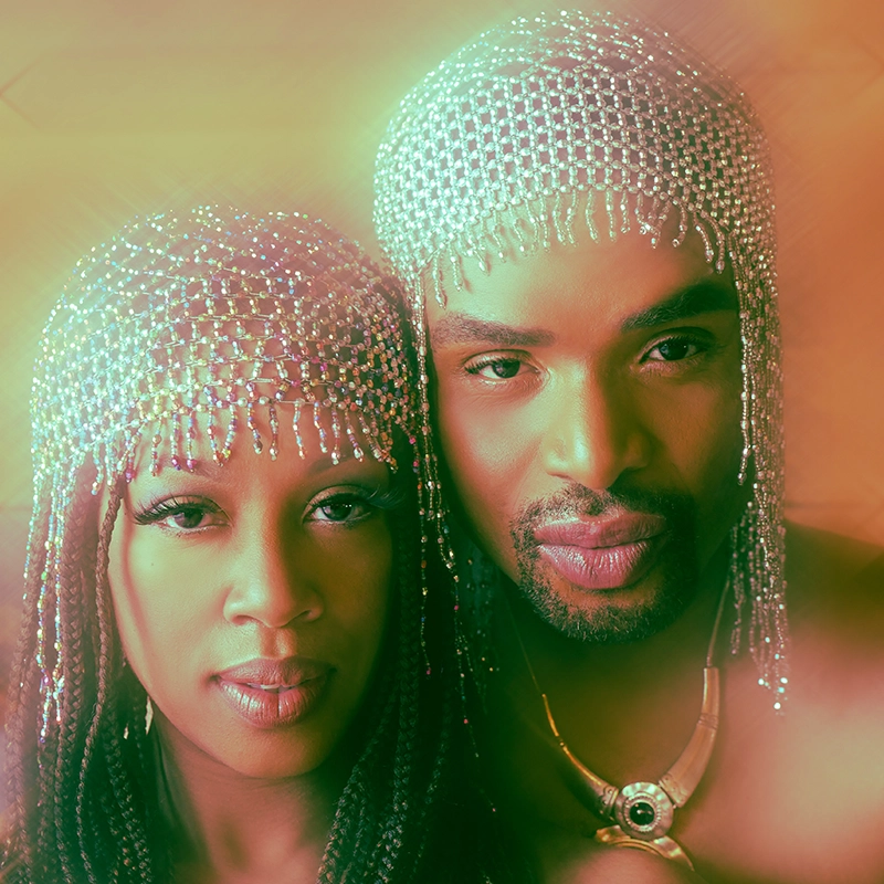 Two people with braided hair wear beaded, shimmering headpieces. Both have serene expressions and are close together, resembling characters from The Incredible Drunkertons. Warm golden light gives the scene a soft, ethereal, and artistic glow.