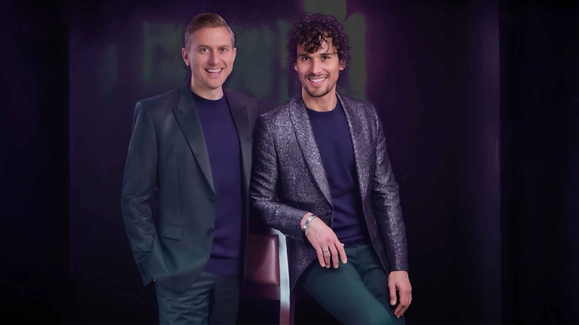 Two men of the piano duo Tiano pose together against a dark, purple-toned background. Both wear dark green pants and jackets over navy shirts. One stands with hands in pockets, smiling; the other, a tenor, sits on a high chair, smiling warmly.