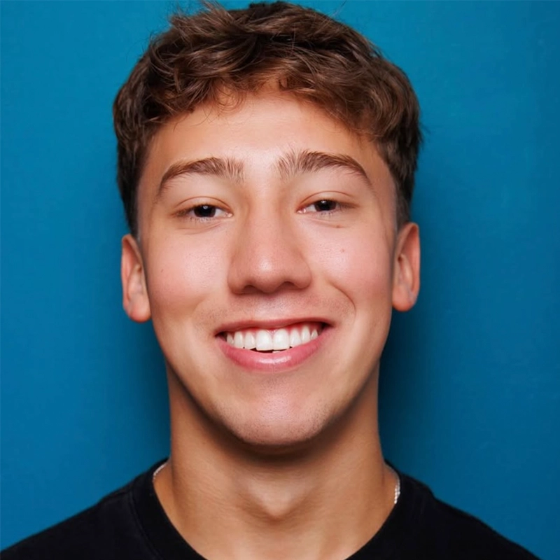 A young man with short, wavy brown hair is smiling broadly, showing his teeth. Wearing a black shirt, Addison Zacharias is positioned in front of a solid blue background, with even lighting highlighting his facial features.