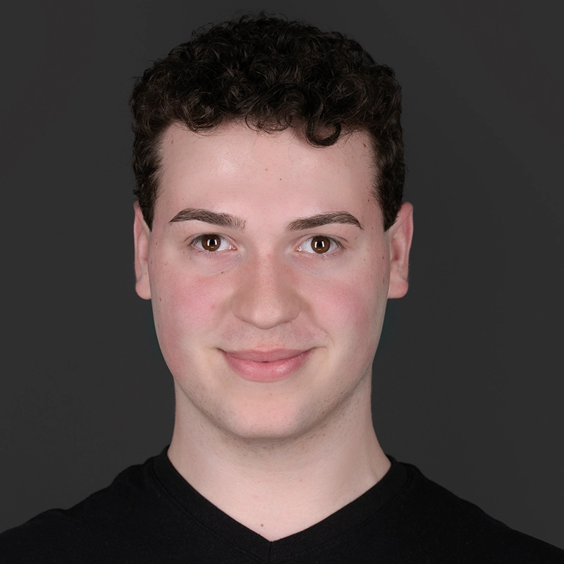 A young person with short, curly dark hair smiles slightly at the camera. They wear a black V-neck shirt and have light skin. The background is solid dark gray, and the lighting is even, emphasizing their facial features.