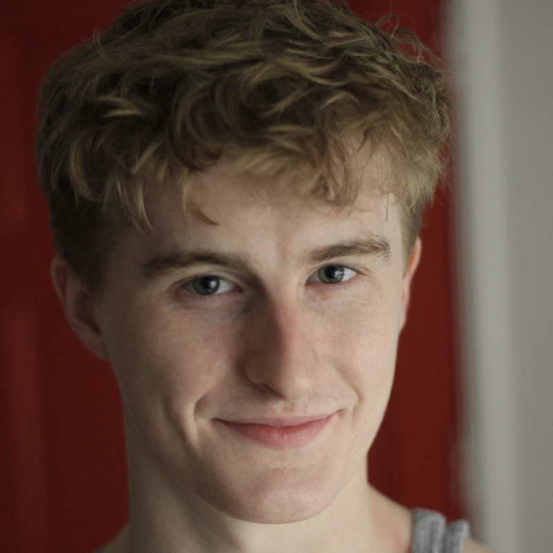 A young person with light skin, wavy blonde hair, and blue eyes smiles softly at the camera. Wearing a gray tank top, they pose against a blurred deep red and white background. The soft natural lighting adds warmth to this Kaylor Toronto portrait.