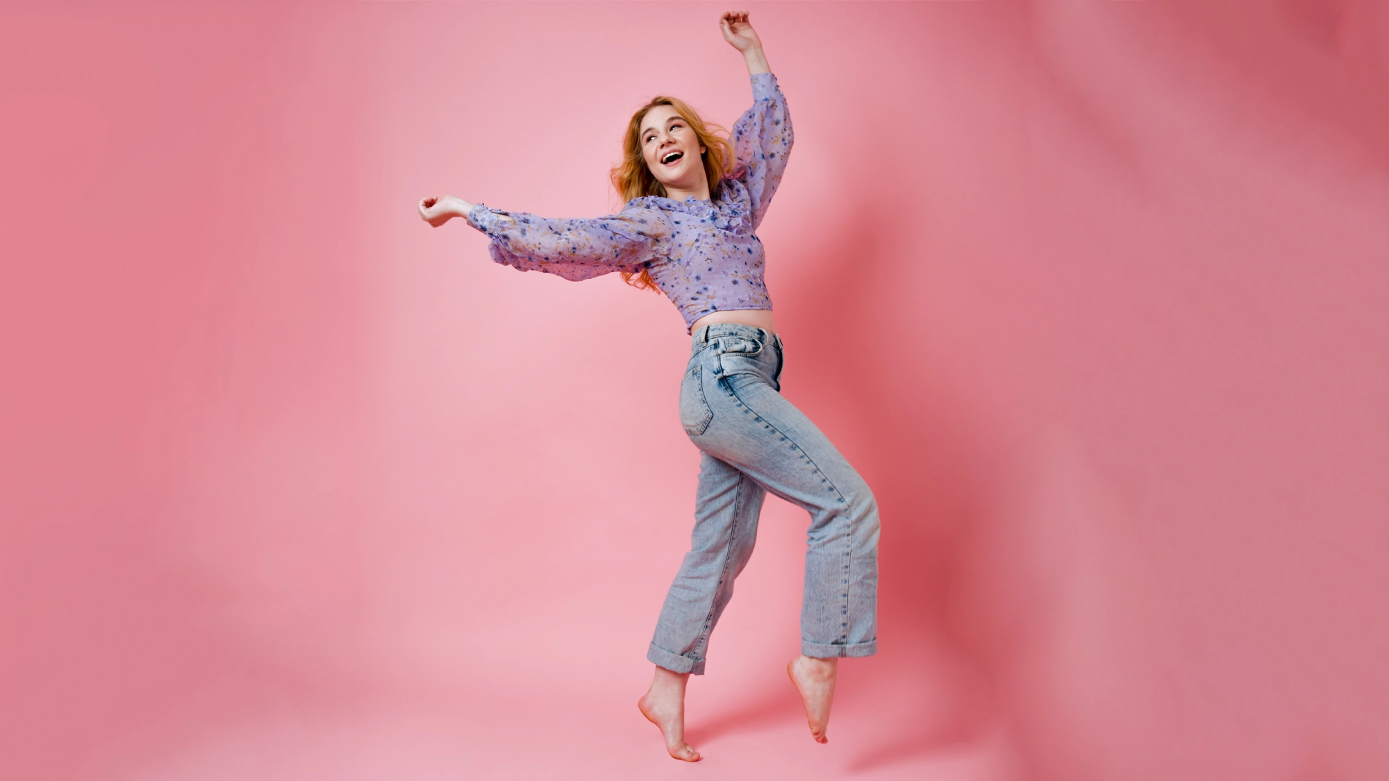 A young woman with wavy red-blonde hair is joyfully jumping against a solid pink background, celebrating her Birthday Extravaganza. She smiles widely in a sheer lavender blouse and light blue high-waisted jeans, arms raised and feet off the ground.
