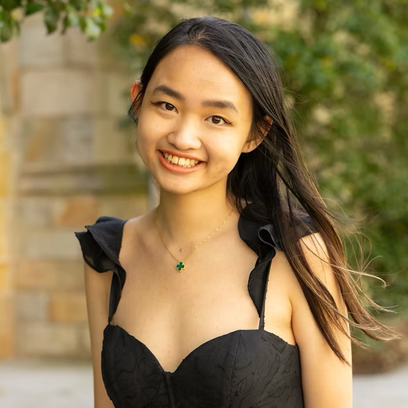 A young woman with long dark hair smiles outdoors. She wears a black sleeveless dress with ruffled straps and a gold necklace with a green four-leaf clover pendant. Sunlight highlights her hair; green foliage and a stone wall are blurred in the background.