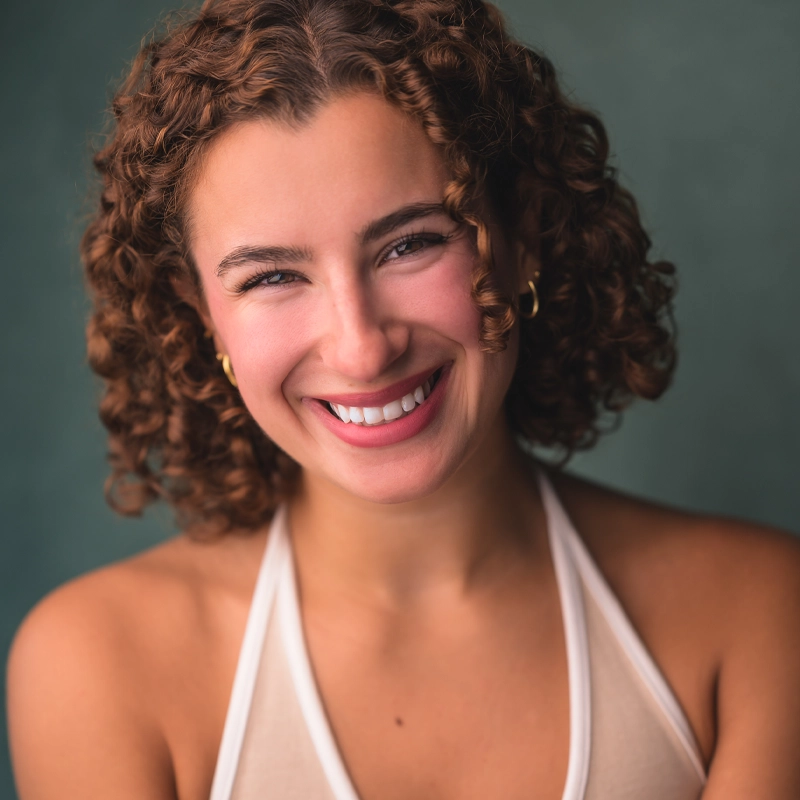 A young woman with short, curly brown hair smiles warmly at the camera. Lexis Trechak wears gold hoop earrings and a cream halter top. The background is a soft, muted green, creating a gentle and inviting atmosphere. Her teeth are visible in her friendly smile.