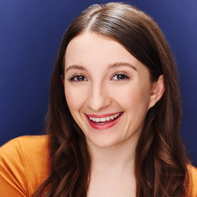 Lexis Trechak, a young woman with long brown hair, smiles brightly at the camera. She wears a mustard yellow top and subtle makeup with pink lipstick against a solid deep blue background, making her outfit and complexion pop.