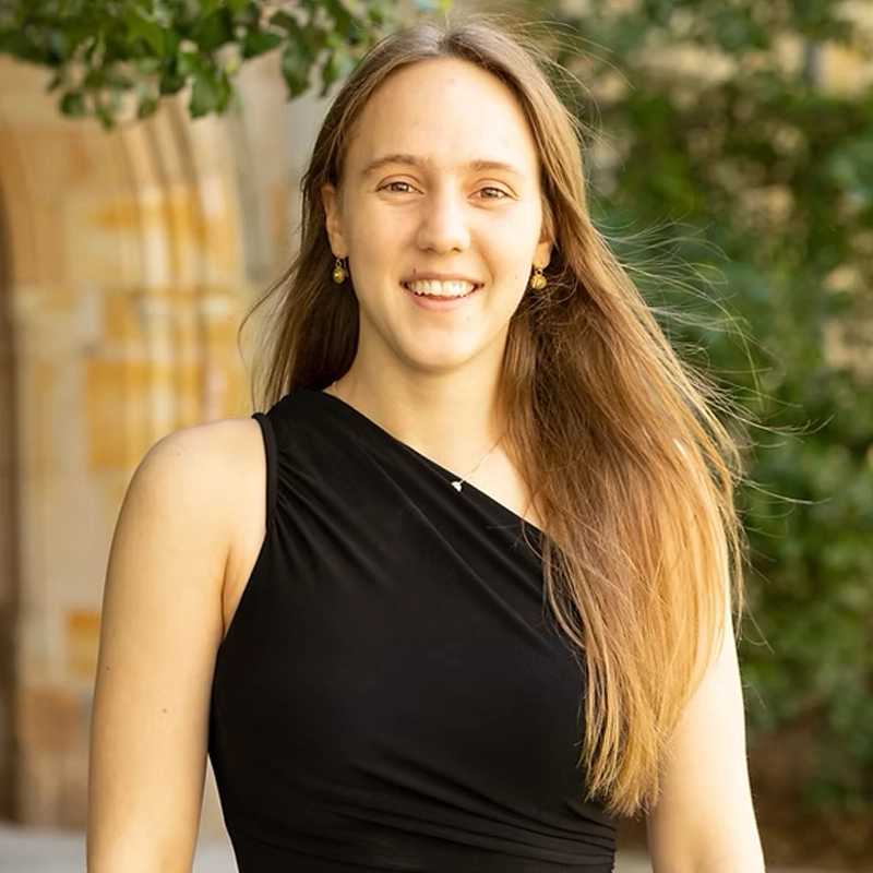 Kate Van Tassel, a young woman with long, straight light brown hair, stands outdoors smiling at the camera in a sleeveless black asymmetrical dress. Green foliage and a stone building with an archway are blurred in the background.
