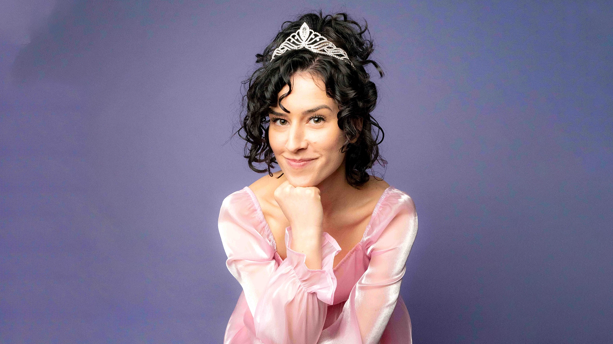 A woman with curly dark hair, resembling a Professional Princess, wears a silver tiara and sheer pink top. She rests her chin on her hands, smiling gently. Against a soft purple backdrop, the dreamy scene hints at charming confessions.