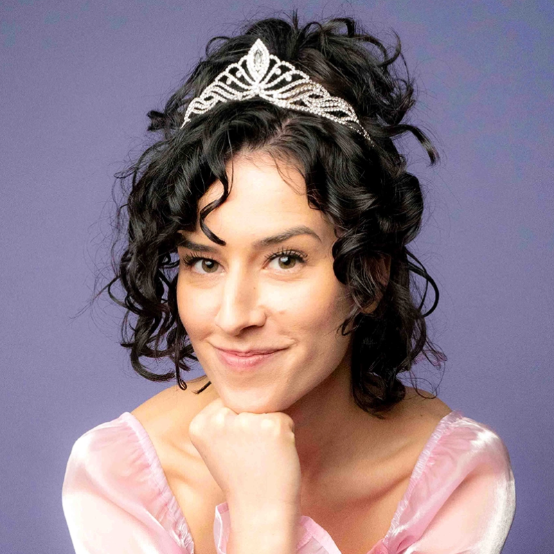 A woman with short curly dark hair wearing a sparkling tiara and a light pink, sheer, off-shoulder top smiles softly. She rests her chin on her hand against a plain purple background.