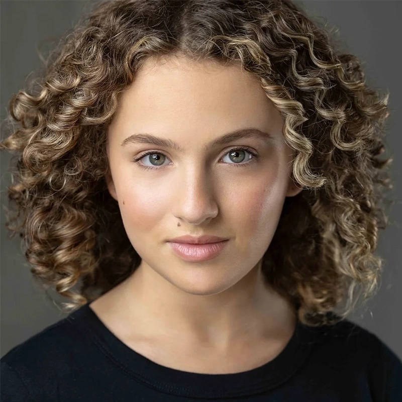 A young person with light skin and expressive green eyes faces the camera, wearing a black top. Violet Tinnirello has short, voluminous, light brown curly hair and a calm, confident expression against a neutral gray background.