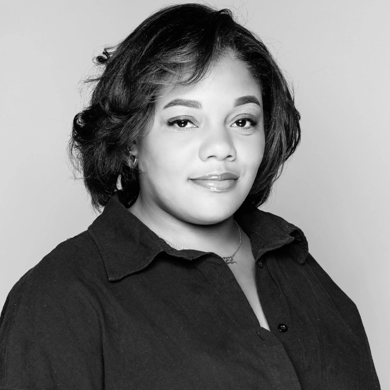 Black and white portrait of a woman with medium-length dark hair styled in waves, wearing a dark button-up shirt. She faces the camera with a slight smile and a confident, calm expression against a plain light background.