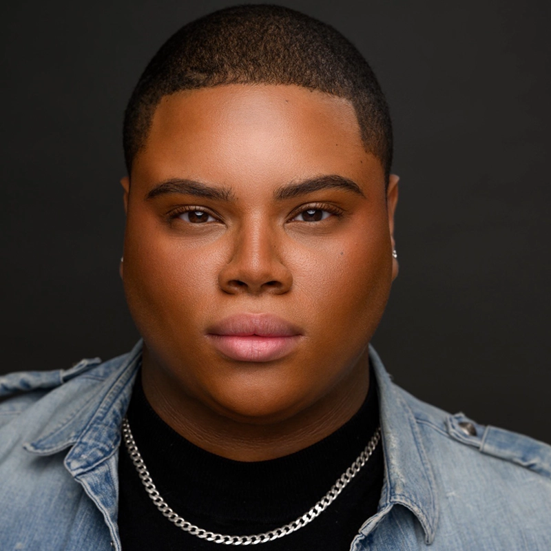 Julian Walton, with short-cropped hair, gazes confidently at the camera. They wear a black shirt, light denim jacket, and a silver chain necklace. Smooth skin and subtle makeup complete the look against a dark background.