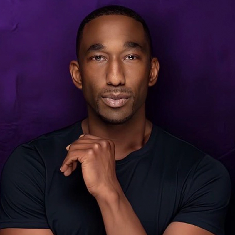Anthony Wayne, with short hair and a trimmed beard, poses against a deep purple background. He wears a fitted black t-shirt and gazes calmly at the camera, resting one hand near his chin in a thoughtful gesture.