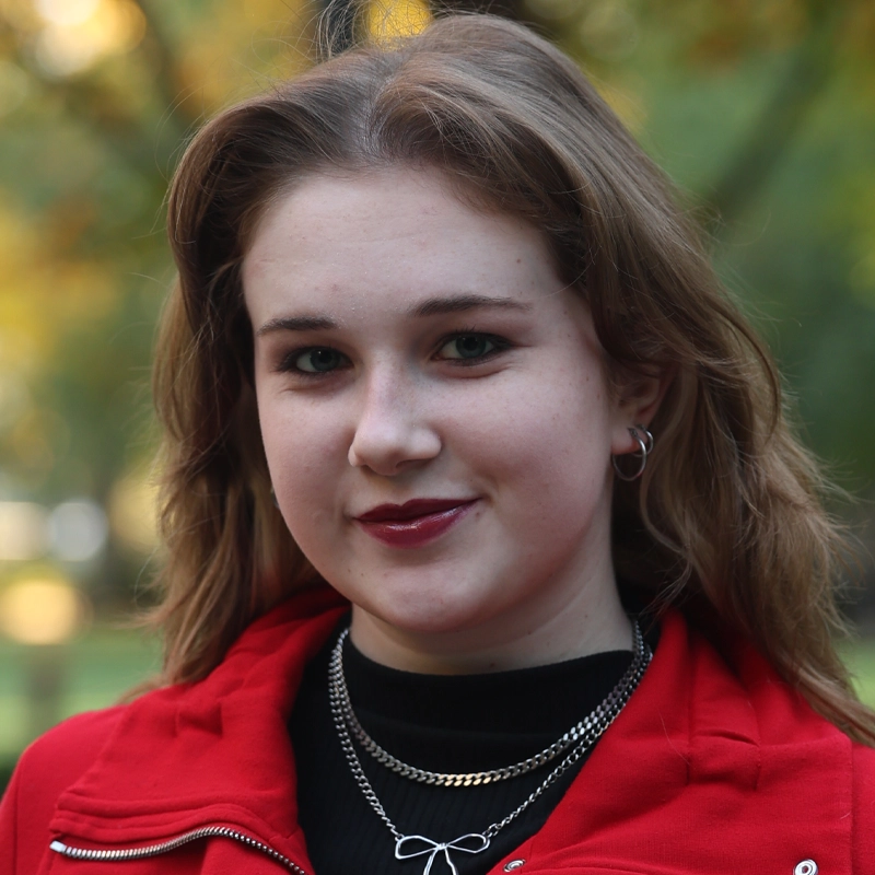 Abbi Wessler, a young woman with light skin and shoulder-length brown hair, smiles softly. She wears a red jacket, black top, layered silver necklaces, and hoop earrings against a backdrop of blurred outdoor greenery.