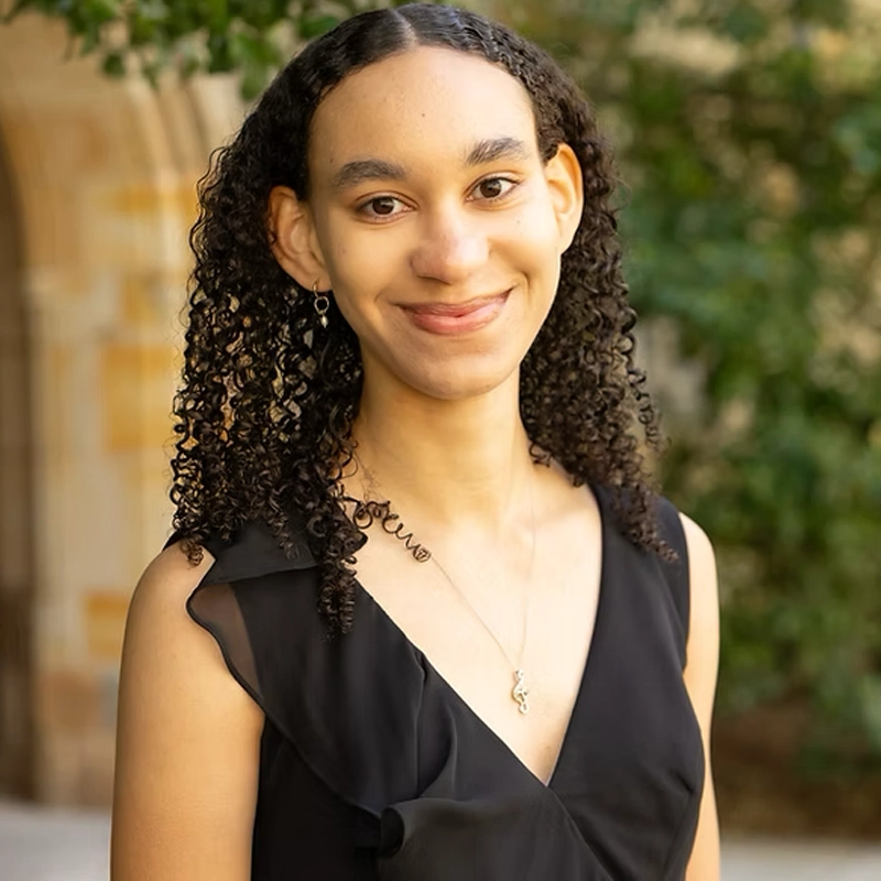 A young person with medium skin and curly dark hair smiles gently outdoors. They wear a sleeveless black dress, a gold necklace, and small hoop earrings. The background is blurred greenery and a stone building, creating a warm, natural setting.