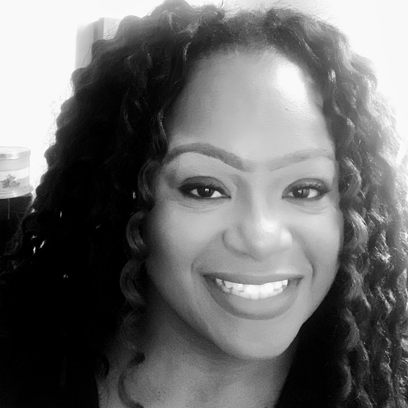 Black and white close-up of Cherette White, a smiling woman with long, curly hair. She has defined eyebrows, wears makeup, and is looking directly at the camera. The blurred background highlights her cheerful expression.