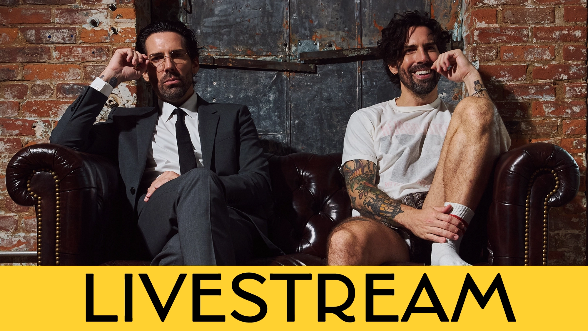 Two men sit on a brown leather couch against a brick wall. One wears a black suit and glasses, the other has tattoos, a white t-shirt, and shorts. Both look at the camera. Bottom yellow banner reads “LIVESTREAM” in bold black letters.