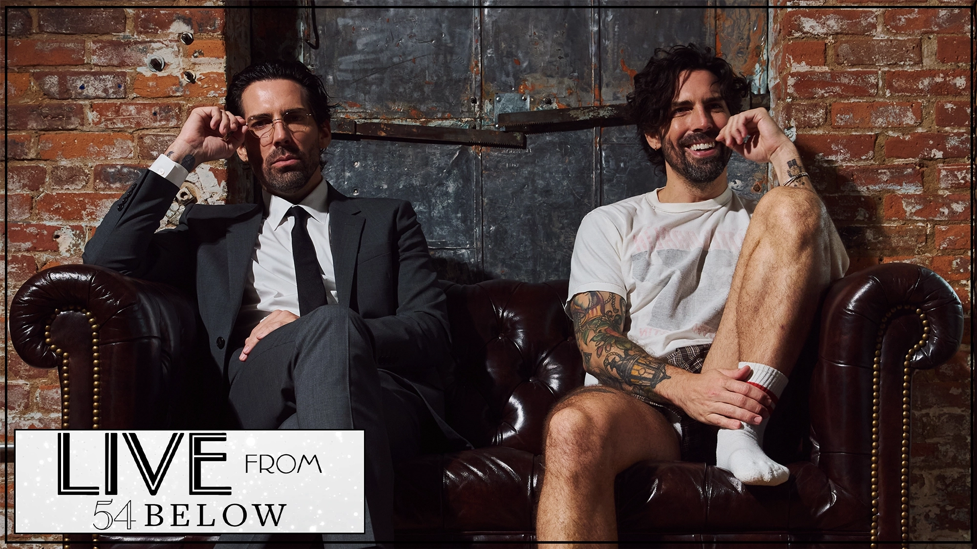 Two men sit on a brown leather couch against an exposed brick wall. The man on the left wears a dark suit and glasses, looking serious. The man on the right is casually dressed, smiling, with tattoos, shorts, and white socks. Text reads “LIVE FROM 54 BELOW.”.