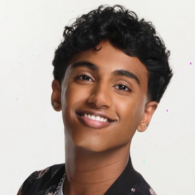 William Alexander, a young person with short, curly black hair, smiles at the camera. They have medium-brown skin and dark eyes, wearing a black top with a faint floral pattern against a plain white background.