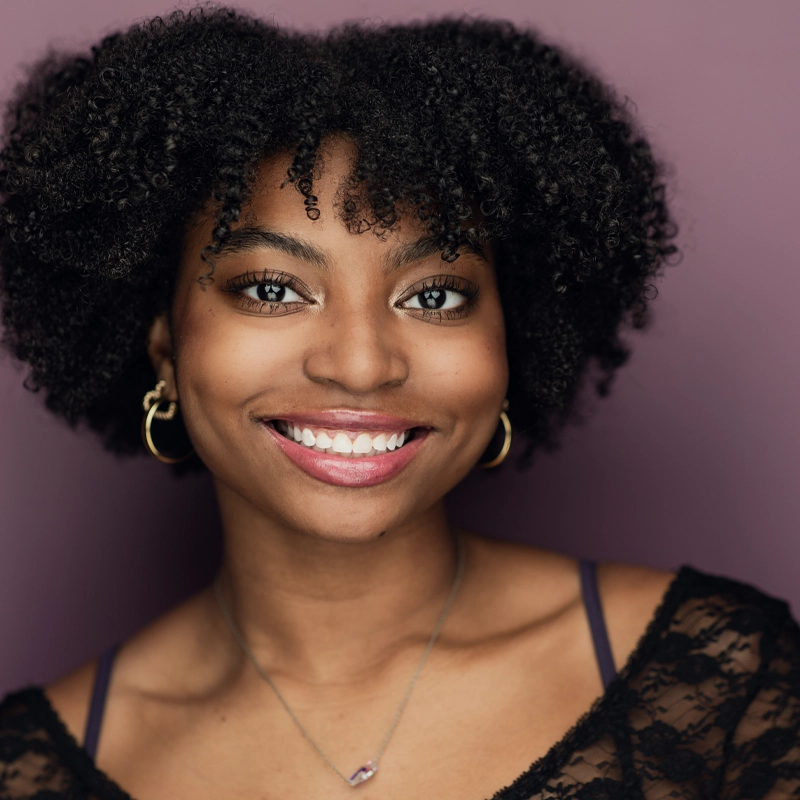 Kendal Williams, a young woman with natural curly hair, smiles warmly at the camera. She wears gold hoop earrings, a delicate necklace, and a black lace top against a solid purple background. Her makeup is natural with pink lipstick.