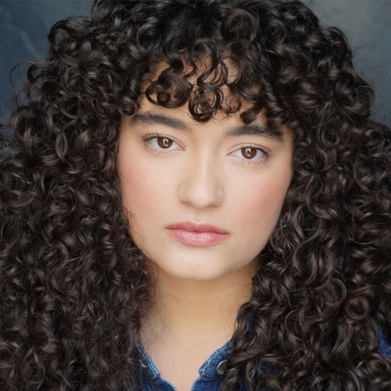 A young person with light skin and long, thick, dark curly hair with short bangs looks directly at the camera. They have brown eyes, full lips, and a neutral expression. The background is dark and slightly blurred. This is Micah Wolfensohn.