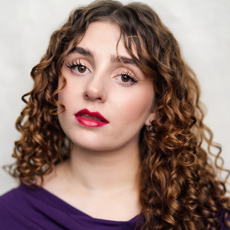 A young woman with long, curly brown hair and light skin looks directly at the camera. She has defined eyebrows, long eyelashes, and wears bold red lipstick. She is wearing a purple top and is posed against a plain, light background.