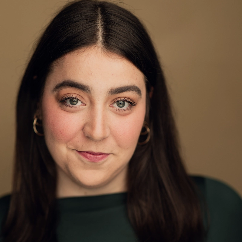 Morgan Zeidman, a woman with long dark hair and fair skin, smiles softly at the camera. She wears gold hoop earrings, subtle makeup with pink blush, and a dark green top against a warm beige background for a polished headshot.