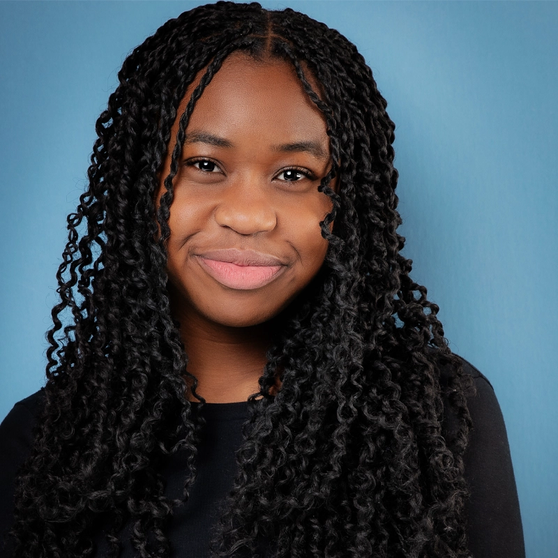 Omolola Adewale, a young woman with long, curly black braids, smiles softly in a black top against a solid light blue background. Her warm, confident expression shines as she faces the camera directly.