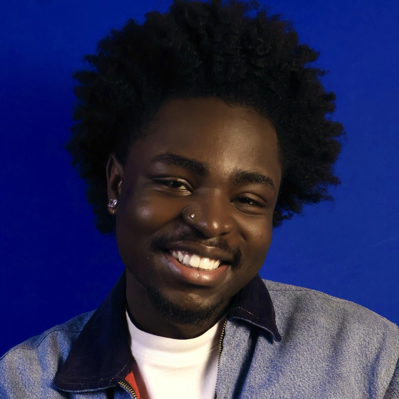 Junior Adjei, a young man with dark skin, short curly hair, and a nose ring, smiles warmly. He wears stud earrings, a light denim jacket with a dark collar, and a white shirt against a solid bright blue background.