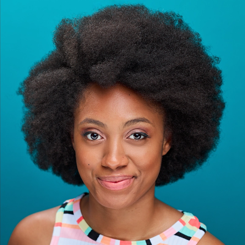 Grace Whetstone smiles gently at the camera, her large, natural afro hairstyle complemented by a pastel, patterned sleeveless top. The solid teal blue background and her soft, natural makeup highlight her radiant presence.