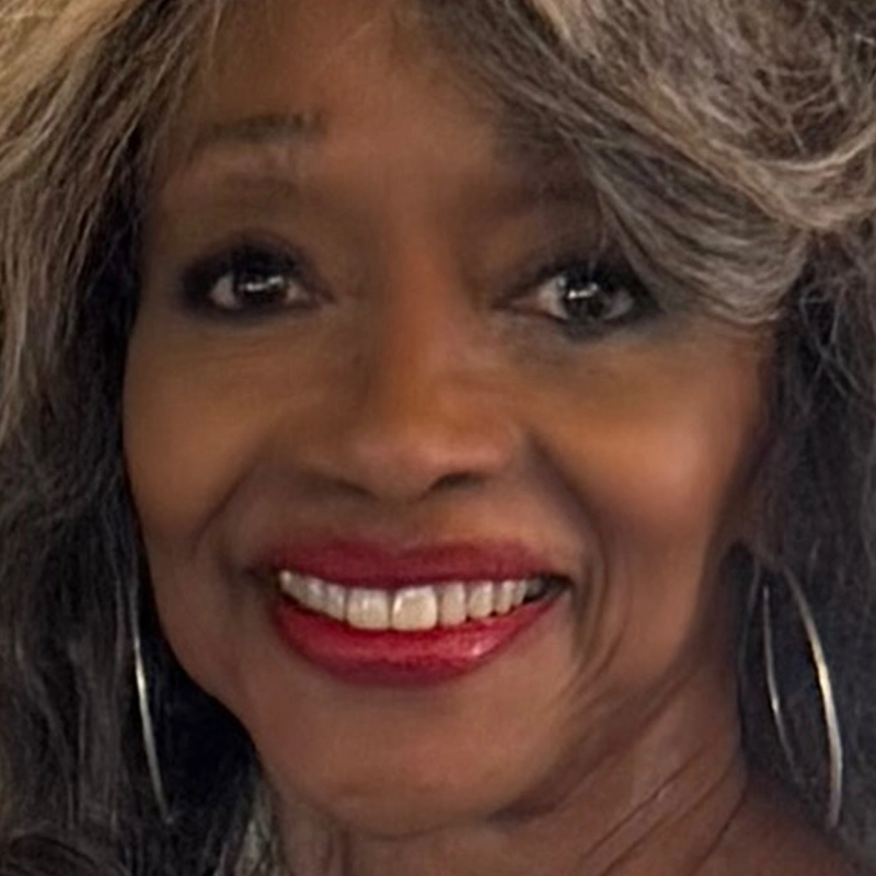 A smiling older woman with long, wavy gray hair, wearing large hoop earrings and bright red lipstick. Her warm, friendly expression and smooth skin resemble Jonelle Allen, while the close-up highlights her prominent cheekbones.