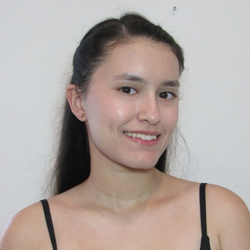 Allison Cento, a young woman with long dark hair, smiles gently at the camera. She wears a black spaghetti strap top and small stud earrings. The background is a plain, light-colored wall with soft, natural lighting.