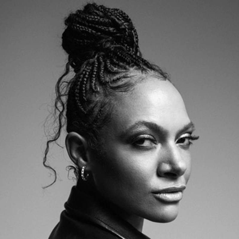 Allison Russell, a Black woman with braided hair styled in a high bun, looks over her shoulder. She has defined eyebrows, long lashes, and wears hoop earrings. The black-and-white image captures her calm, confident expression.