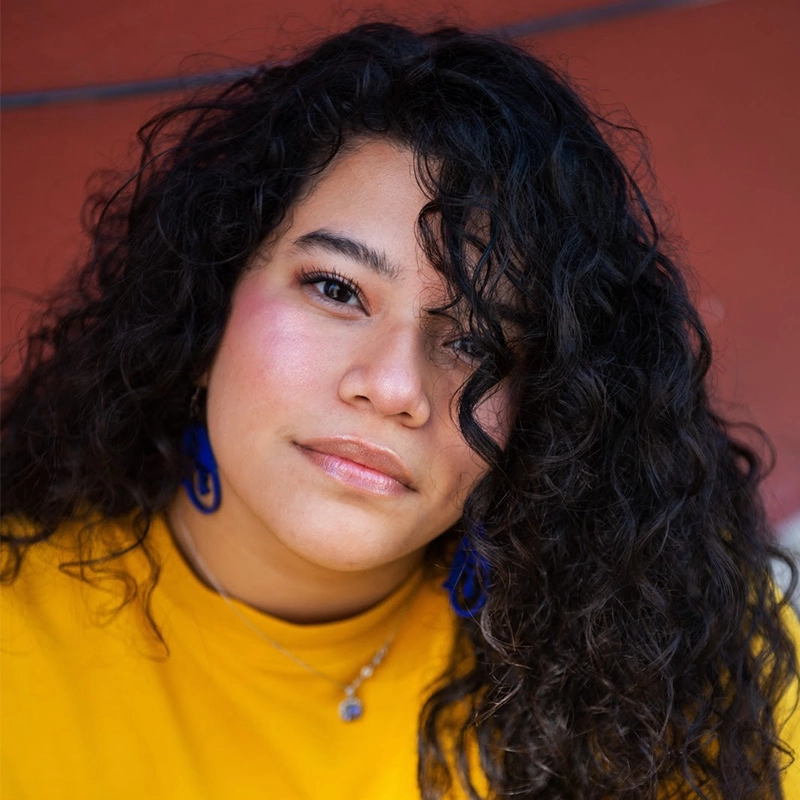 Grace Whetstone, with long, curly dark hair and glowing skin, gazes at the camera. She wears a yellow top, blue statement earrings, a necklace, and natural makeup with rosy cheeks. The blurred background is reddish-brown.