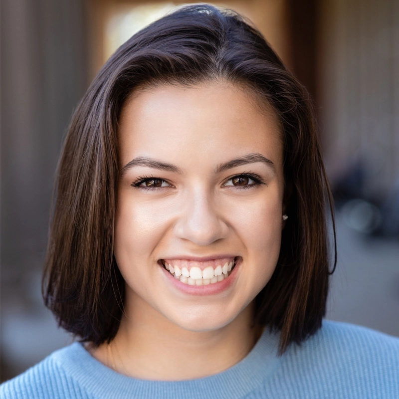 A young woman with straight, shoulder-length brown hair smiles warmly at the camera. She has brown eyes, even features, and wears a light blue sweater. The softly blurred background hints at an outdoor setting captured by Spencer Lawson.