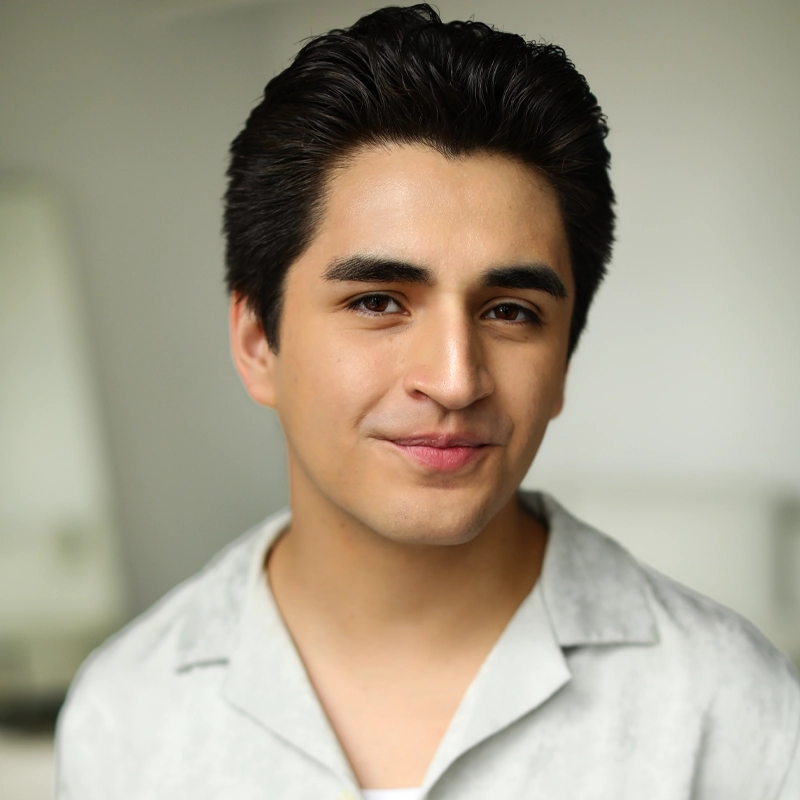 A young man with dark hair, styled neatly, smiles softly at the camera. He has fair skin and is wearing a light gray collared shirt over a white top, reminiscent of Marcus Alonso’s classic style. The blurred background suggests an indoor setting.