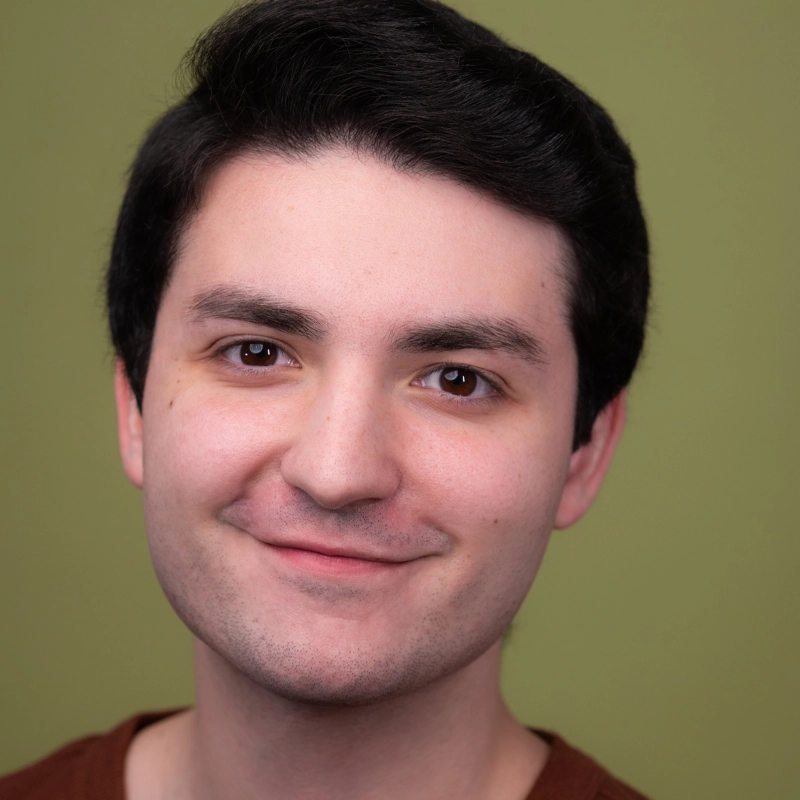 John Amalfitano, a young man with short dark hair and fair skin, is smiling slightly at the camera. He wears a brown shirt and is posed before a plain olive green background, with even lighting highlighting his facial features clearly.