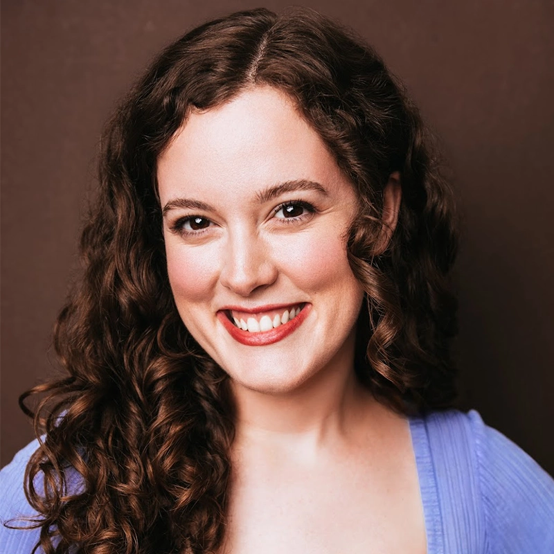 Madison Keenoy, with long, dark curly hair, smiles warmly at the camera. She wears a light purple top with a deep neckline and has a light complexion. The soft lighting highlights her face and hair against the solid brown background.