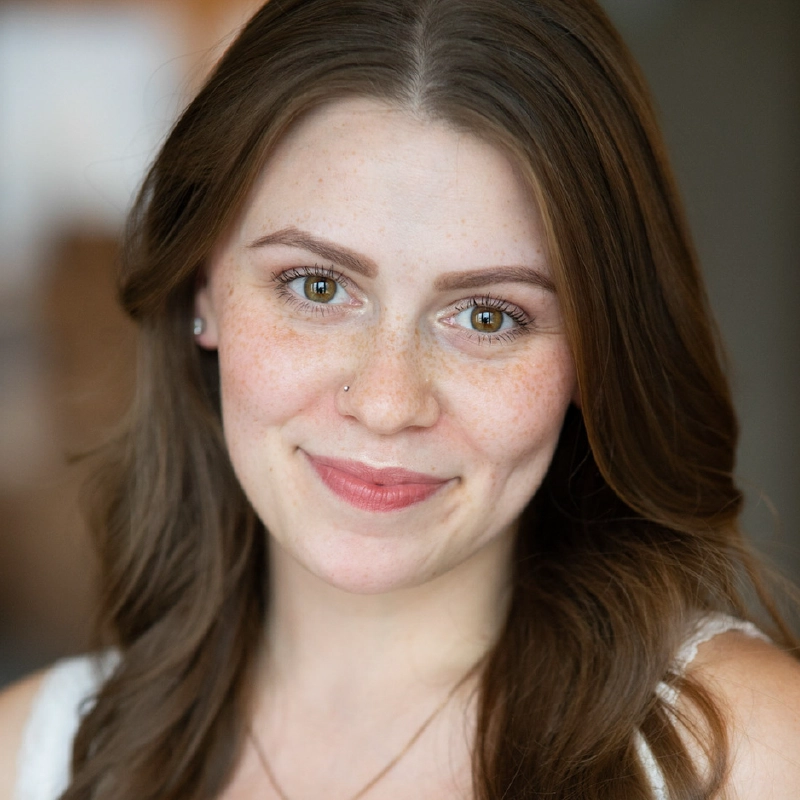 A young woman with long brown hair and light skin smiles softly. She has green eyes, light makeup, freckles, a nose piercing, and small earrings. Inspired by the natural style of Gabe Woodard, she wears a white top against a warmly blurred background.