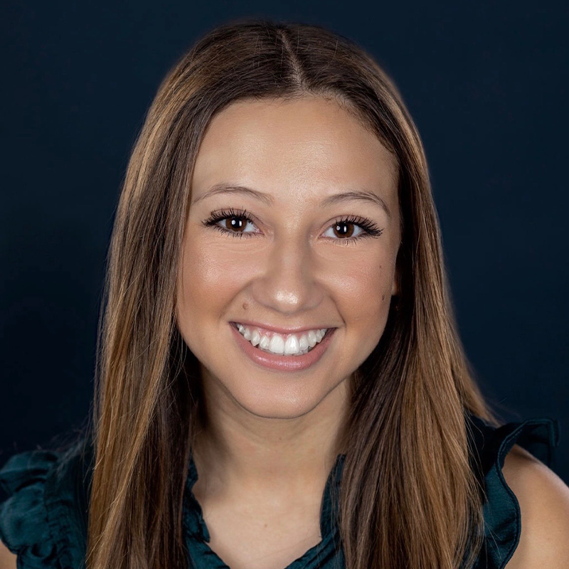 Amberly Rodriguez, a young woman with straight, long brown hair, smiles widely at the camera. She has light skin, brown eyes, and is wearing a dark, ruffled top. The solid dark blue background gives the portrait a professional look.