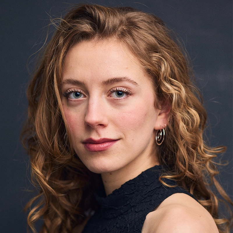 A young woman with wavy, light brown hair and blue eyes looks at the camera. She is wearing a sleeveless black top with a lace neckline and small hoop earrings. The background is dark and plain, highlighting her natural features.