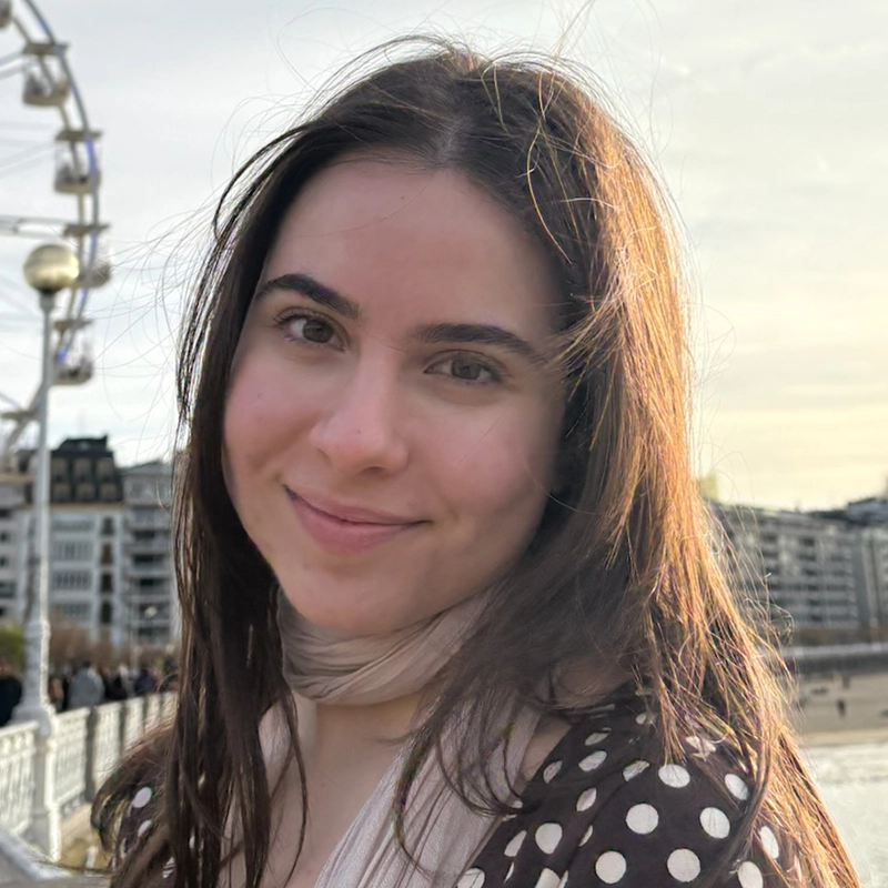 A young woman with long brown hair smiles gently at the camera. She wears a dark top with white polka dots and a light scarf. The background shows buildings, a ferris wheel, and a fence on a sunny day, with soft sunlight lighting her hair.