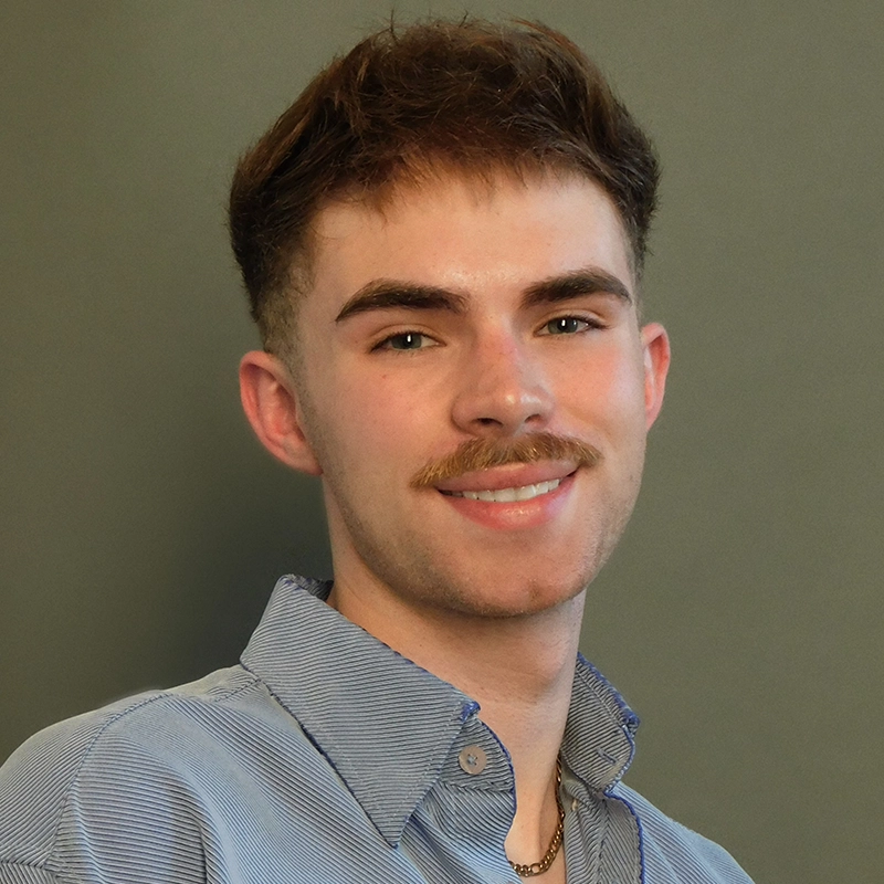 Gabe Woodard, a young man with short brown hair and a trimmed mustache, smiles at the camera. He is wearing a light blue collared shirt over a gold chain necklace, set against a plain, muted olive green background.