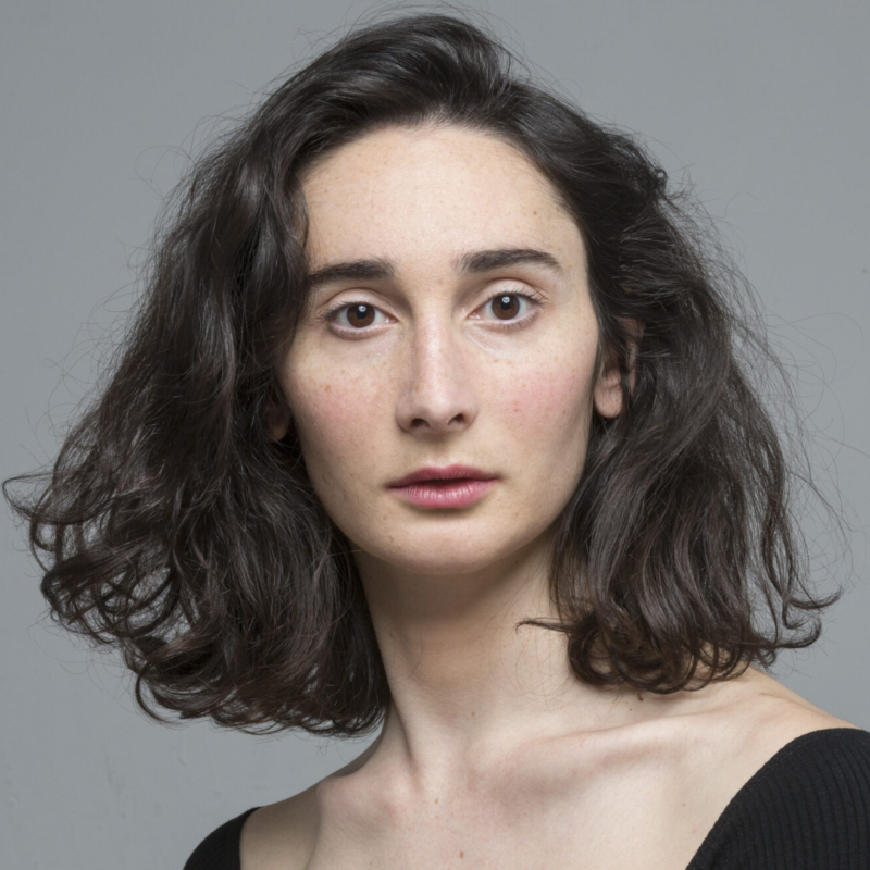 A woman with pale skin, wavy shoulder-length dark brown hair, and brown eyes stares seriously at the camera. She wears a black top with a wide neckline. The background is plain, light gray, and the lighting is soft and even.